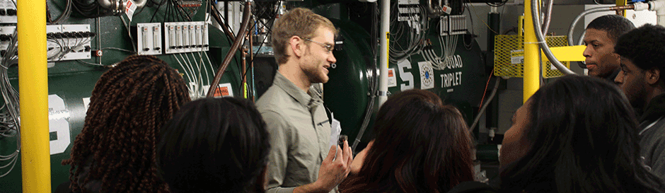 A tour guide explains the cyclotrons to a group of high school students.